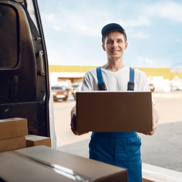 Deliveryman in uniform unloads the car with parcels, delivery service. Man standing at cardboard packages in vehicle, male deliver, courier or shipping job