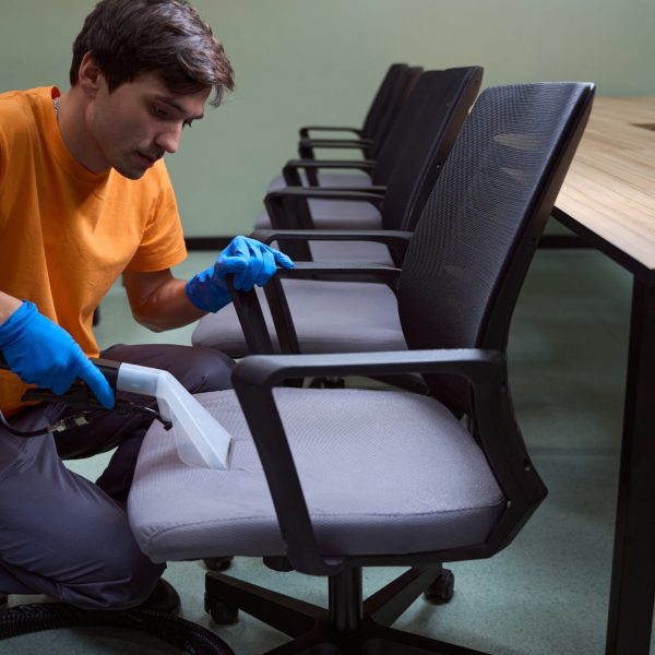 Attentive young man in rubber gloves using an upholstery vacuum-cleaner on office furniture